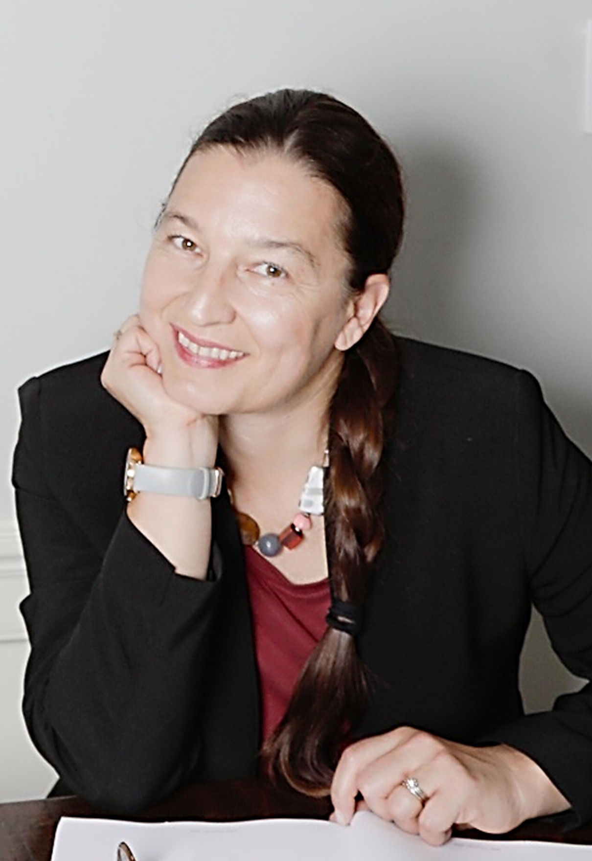 Aleksandra Pikula, MD &mdash; founder of HER PRISM, at her desk in a black blazer with warm smile and chin resting on hand